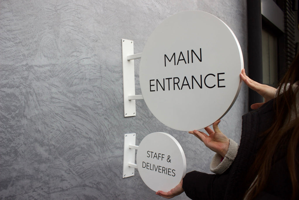 Twee ronde witte aluminium uithangborden als bewegwijzering bij een ingang
